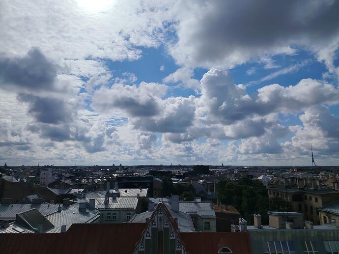 Panorāmas skats uz Rīgas pilsētas jumtiem zem mākoņainām debesīm, caur kurām spīd saule. (Captioned by AI)