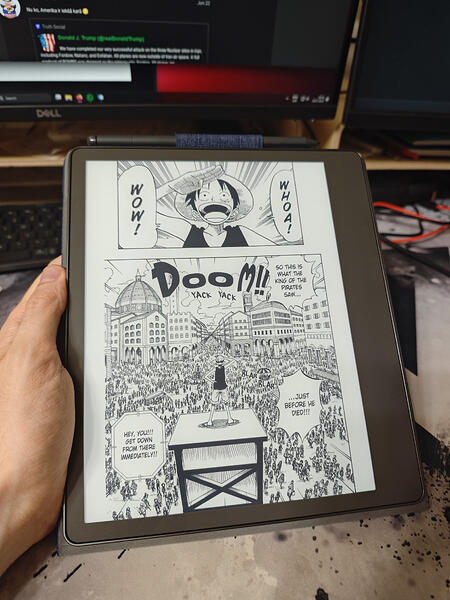 Cilvēks tur planšetdatoru ar atvērtu manga lapu, fonā redzams datorekrāns. (Captioned by AI)