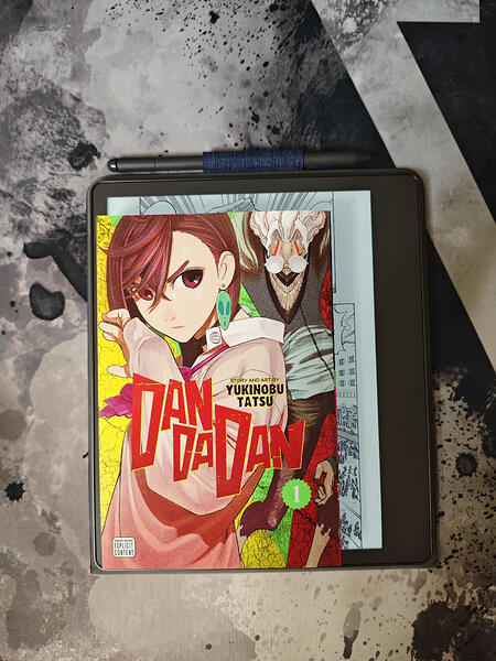 Manga "Dandadan" pirmais sējums novietots uz melna e-lasītāja, ar irbuli virs tā. (Captioned by AI)