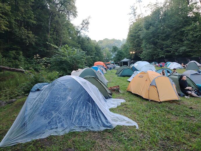 Kempinga laukums, kurā ir daudz telšu uz zālāja starp kokiem, un atpūšas cilvēki. (Captioned by AI)